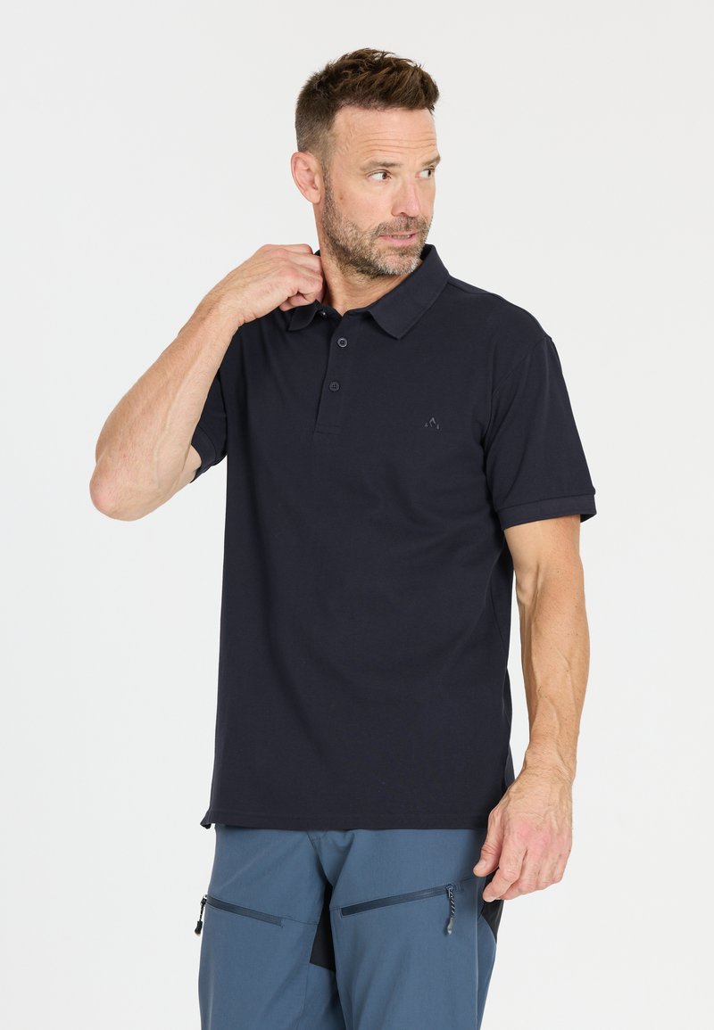 Man die een zwart poloshirt met korte mouwen en een blauwe broek draagt, zijn kraag aanraakt en naar links kijkt tegen een effen lichte achtergrond.