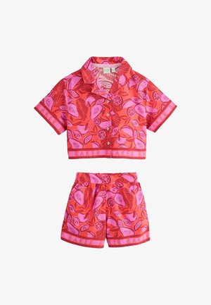O fată tânără poartă o cămașă cu mâneci scurte, imprimeu tropical roșu și roz, și pantaloni scurți asortați cu sandale albe, stând pe un fundal simplu.