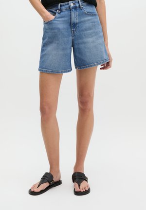 Frau trägt mittelblaue Jeansshorts mit Seitentaschen und schwarzen Slip-On-Sandalen, steht vor einem einfarbigen Hintergrund.