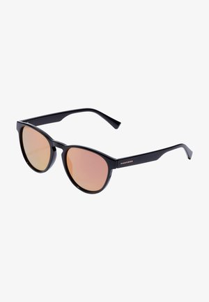 Lunettes de soleil noires en plastique avec des montures rondes et des verres teintés rose. Elles présentent des branches fines avec le logo "HAWKERS" en détail métallique.