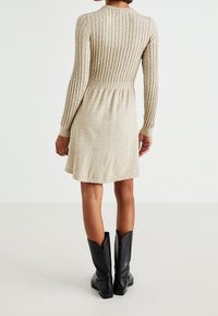 Femme portant une robe beige en tricot à manches longues et des bottes en cuir noires mi-mollet, debout, dos au spectateur.