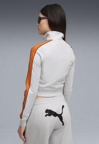 Witte cropped sweatshirt met hoge kraag, oranje zijstrepen en zwarte accenten; gecombineerd met lichtgrijze broek met een zwart Puma-logo.