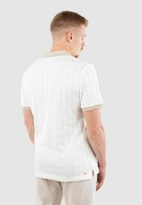 Polo en tricot blanc à manches courtes, avec une texture quadrillée et des accents beiges au niveau du col et des bords des manches.