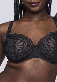 Reggiseno in pizzo nero con coppe trasparenti e ricami floreali, spalline regolabili e un accento dorato al centro. Design semplice ma elegante.