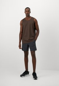 Brun ærmeløs tanktop med rund halsudskæring, parret med mørkegrå shorts; modellen bærer sorte træningssko. Ensfarvet baggrund.