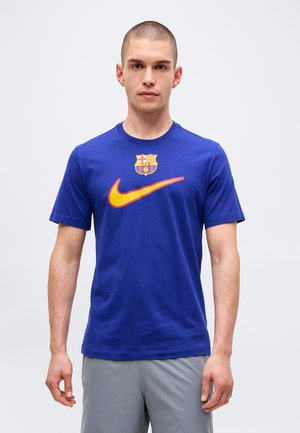 Homme debout face à l'avant portant un t-shirt bleu avec un swoosh Nike orange et le blason du FC Barcelone sur la poitrine, short gris.
