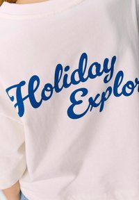 Camiseta de algodón blanca con corte holgado. Presenta un texto en cursiva azul que dice "Explorador de Vacaciones" en la parte de atrás. Diseño minimalista.