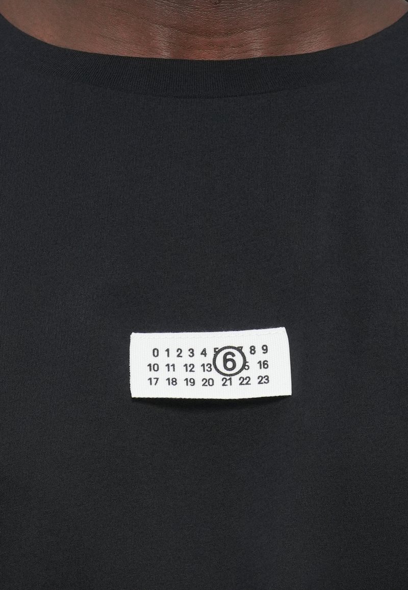 Sort bomuld t-shirt med et hvidt stofmærke med trykte numre og et cirkulært ikon, placeret på venstre brystområde.