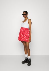 Top branco sem mangas emparelhado com uma saia vermelha com padrão floral. Modelo usa óculos de sol pretos, botas pretas e meias brancas. Design suave e casual.