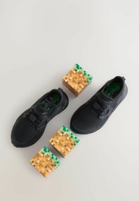 Baskets de sport noires avec un tissu texturé, dotées de semelles intérieures vertes, accompagnées de blocs pixelisés ressemblant à des blocs de terre d'un jeu vidéo.