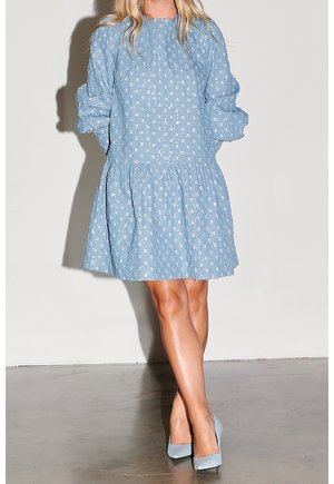 Kobieta ubrana w jasnoniebieską sukienkę z długim rękawem w biały kwiatowy wzór oraz pasujące niebieskie szpilki, stojąca ze skrzyżowanymi nogami na szarej podłodze.