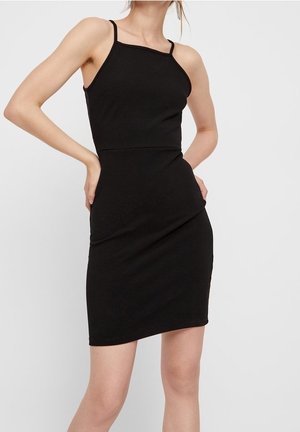 Kobieta ubrana w dopasowaną, czarną mini sukienkę bez rękawów z cienkimi ramiączkami, stojąca z rękami na biodrach na tle jednolitej białej ściany.