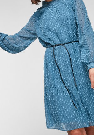 Kobieta w długiej sukience z teksturowanego niebieskiego materiału, z cienkim czarnym paskiem luźno związanym w talii, na jednolitym tle.