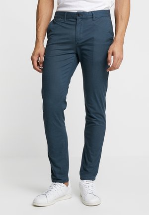 Mann trägt dunkelblaue eng anliegende Hose und weiße Sneaker, steht vor einfachem hellem Hintergrund.