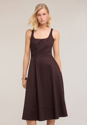 Mujer rubia con vestido midi sin mangas de color marrón oscuro con escote cuadrado, pendientes de aro dorados y pulseras doradas sobre fondo liso.