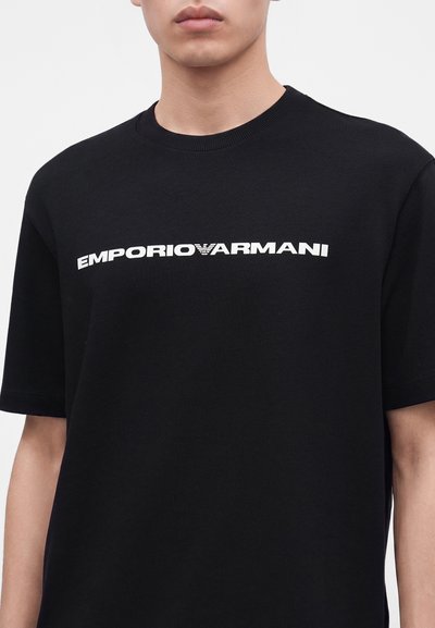 Νεαρό άτομο που φοράει μαύρο T-shirt Emporio Armani με λευκό λογότυπο στο στήθος, εμφανίζεται από τον λαιμό έως το μέσο του κορμού.