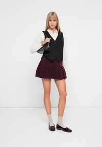 Zwarte mouwloze vest, witte blouse en kastanjebruine geplooide rok. Beige sokken en schoenen van gepatenteerd leer in het donker completeren de outfit. Eenvoudig, strak ontwerp.