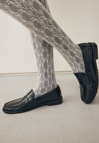 Mocassins en cuir noir à finition lisse avec une bride décorative, associés à des collants gris à motifs floraux.