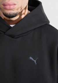 Sort hoodie med hætte, lavet af blødt stof. Har et gråt broderet Puma-logo på brystet. Glat tekstur, enkelt design.