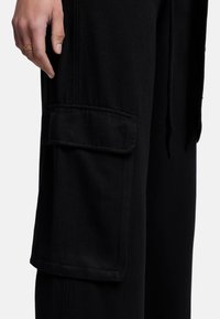 Schwarze Cargo-Hosen aus einem weichen, strukturierten Stoff. Sie verfügen über eine große, flache Cargo-Tasche an der Seite und einen Kordelzug in der Taille.