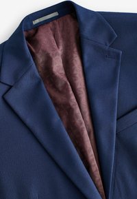 Marineblazer mit strukturiertem Äußeren, das ein bordeauxfarbenes, gemustertes Futter zeigt, das am Kragen und an den Revers sichtbar ist. Taillierte Form und elegantes Design.