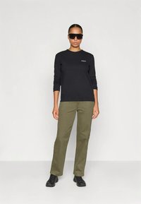 Μαύρο πουκάμισο με μακριά μανίκια και μικρό λογότυπο, παντελόνι σε ελιά πράσινο και μαύρα παπούτσια. Το άτομο φοράει σκουρόχρωμα γυαλιά ηλίου και στέκεται μπροστά σε ουδέτερο φόντο.