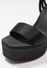 Černé platformové sandály s širokým saténovým popruhem a texturovaným matným podkladem. Obuv má zaoblenou špičku a plochou konstrukci podpatku.