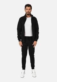 Giacca nera con zip sopra una maglietta bianca, abbinata a pantaloni jogger neri. Vestibilità comoda con polsini elasticizzati e sneakers bianche.