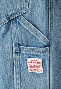 Gaiši zilas denima audums ar lielu kabatu un sarkanu un baltu birku ar tekstu "kad ir jāstrādā, LEVI STRAUSS & CO."