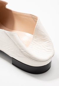 Chaussure blanche en cuir lisse avec un bout arrondi et un petit talon bloc noir. Présente une texture subtile sur la surface supérieure et une doublure intérieure neutre.