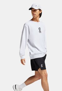Sweatshirt blanc avec logo noir, associé à un short noir avec logo blanc. Une casquette gris clair avec un design perforé complète la tenue.
