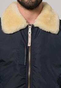 Marinblå bomberjacka med en beige pälsiga krage, metalldragkedja, tygögla och texturerade lappar på framsidan.