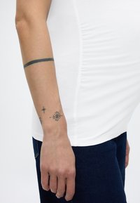 Ruka s crnim kružnim tetovažnim remenom na gornjem dijelu podlaktice i tetovažama zvjezdastog oblika blizu zapešća, odjevena u bijelu majicu i tamnoplave traperice.