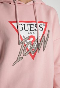 Zbliżenie różowej bluzy z kapturem z logiem GUESS w kolorze czerwonym i białym oraz tekstem "Icon" z koralików na piersi, z różowymi sznurkami ściągającymi zakończonymi metalowymi końcówkami.