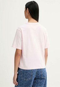 T-shirt di cotone rosa chiaro, vestibilità comoda, maniche corte; indossata con pantaloni di jeans blu con motivo logo su tutta la superficie, vista posteriore.