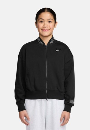 Joven con cabello oscuro con chaqueta negra con cremallera de Nike y pantalones gris claro, de pie frente a un fondo blanco liso, sonriendo.