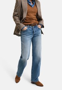 Helle blaue Jeans mit weitem Bein, getragen mit einem braunen Zopfstrickpullover unter einem gemusterten Blazer. Braune Schuhe vervollständigen das Outfit.