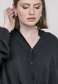 Chemisier noir à boutonnage, en tissu doux, avec un décolleté en V et des manches longues ; il comprend des boutons noirs et une coupe décontractée.