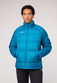 Veste doudoune turquoise avec col montant, fermeture éclair et logo sur la poitrine. Présente une coupe carrée et une texture douce et matelassée.