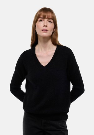 Femme aux cheveux roux lisses portant un pull noir à col en V et un pantalon noir, debout avec les mains derrière le dos sur un fond clair uni.