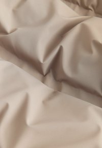 Beige Stoff mit einer glatten, strukturierten Oberfläche, die ausgeprägte Falten und einen subtilen Glanz aufweist. Das Material scheint leicht und flexibel zu sein.