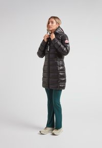 Mujer ajustando el cuello de un abrigo de plumas largo y negro, vistiendo pantalones de color teal y zapatillas beige, de pie frente a un fondo sencillo.