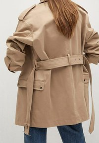 Manteau beige à ceinture et poches à rabat porté sur un jean bleu, vu de dos sur une personne avec de longs cheveux châtain clair.
