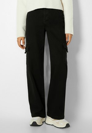 Osoba ubrana w czarne szerokie spodnie typu cargo, biały sweter i białe trampki stojąca na jednolitym tle.