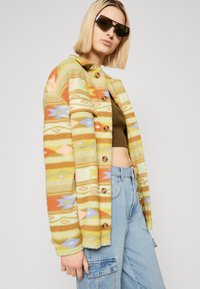Fleece jack met meerkleurige geometrische patronen in oranje, geel en blauw; voorzien van grote knopen en een kraag.