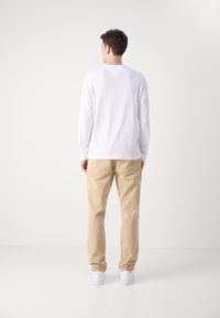 Camisa blanca de manga larga con cuello redondo, combinada con pantalones beige. El atuendo se completa con zapatillas blancas, visto desde la parte de atrás.