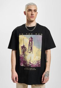 Sort bomuld t-shirt med et farverigt grafik af en raketopsendelse, text nederst, og afslappet pasform. Modellen bærer en sølvkæde.