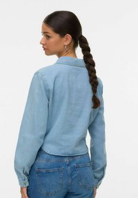 Camicia di jeans azzurro chiaro con maniche lunghe, polsini con bottoni, colletto e orlo curvo. Abbinata a jeans blu, vista da dietro.