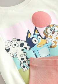 Sudadera de color claro con un estampado gráfico de tres perros de dibujos animados sobre un fondo en tonos pastel; detalle de cinturilla acanalada en rosa.