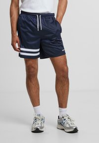 Shorts de sport navy avec des rayures blanches à l'ourlet, une taille élastique avec cordon de serrage et des poches. Portés avec des baskets grises et des chaussettes blanches.